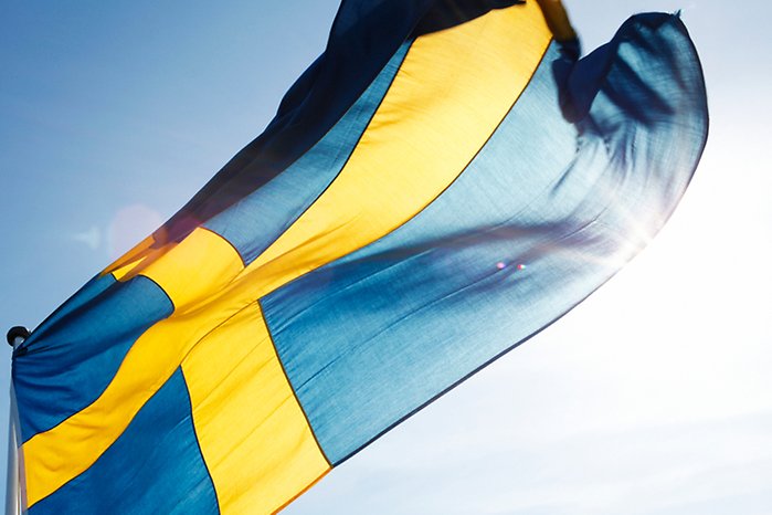 Foto på en vajande svensk flagga med blå botten och gult kors med solen som strilar igenom tyget mot en klarblå himmel. 